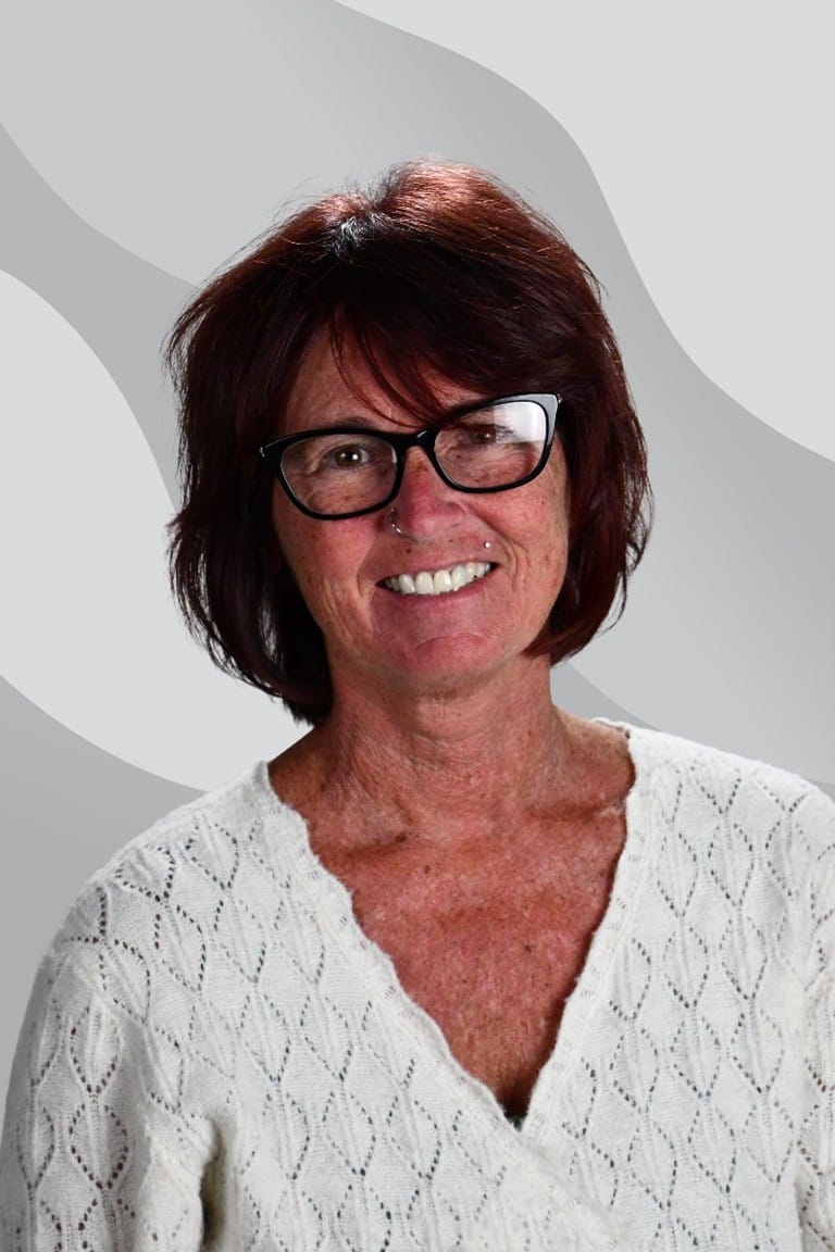 A smiling woman with auburn hair and glasses wearing a white patterned sweater.