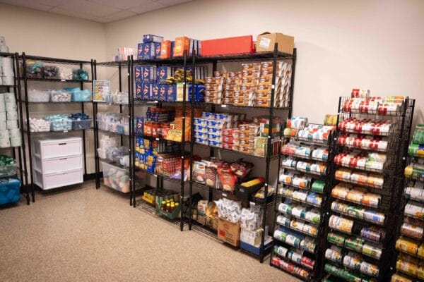 Metal shelving units in a pantry filled with food items including canned goods, boxes of pasta and rice, and small packages of snacks.