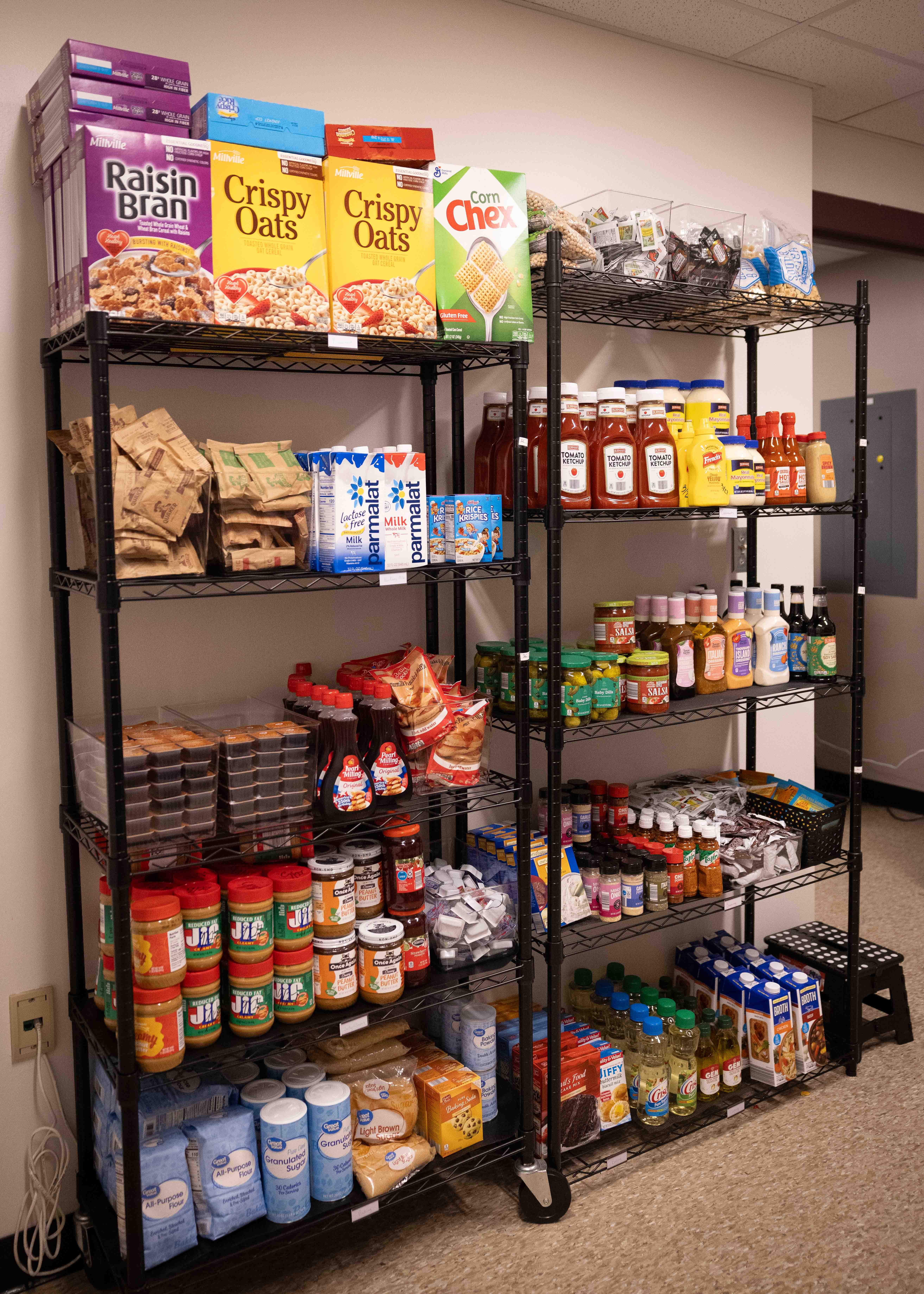 A tall wire shelving unit stocked with a variety of groceries including cereal, jars of peanut butter, condiments, bottles of oil, and bags of flour and sugar.